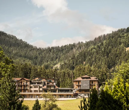 Excelsior Dolomites Life Resort © Hannes Niederkofler Hotelgebäude eingebettet in grüne Berge und Wald unter blauem Himmel