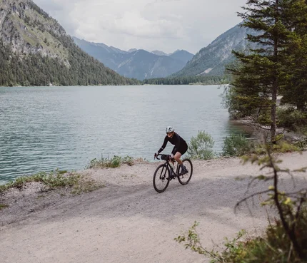 Gravel.Tirol © Liz Kellerer Fahrradfahrer auf Schotterweg neben See und Bergen
