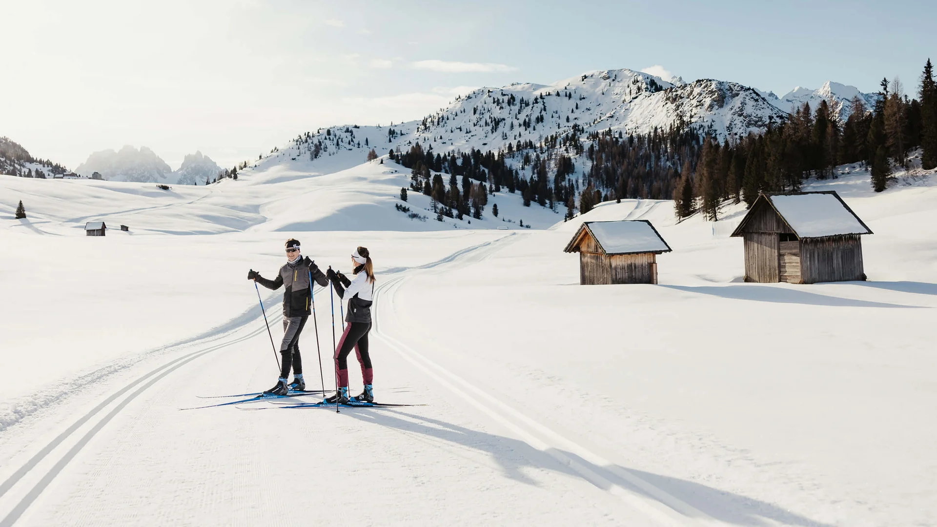 MTS Austria Two cross-country skiers on groomed trail in snowy mountain landscape with huts