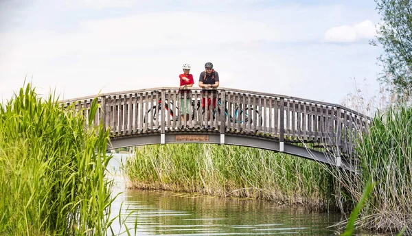 Two cyclists standing on a wooden bridge over a river surrounded by reeds