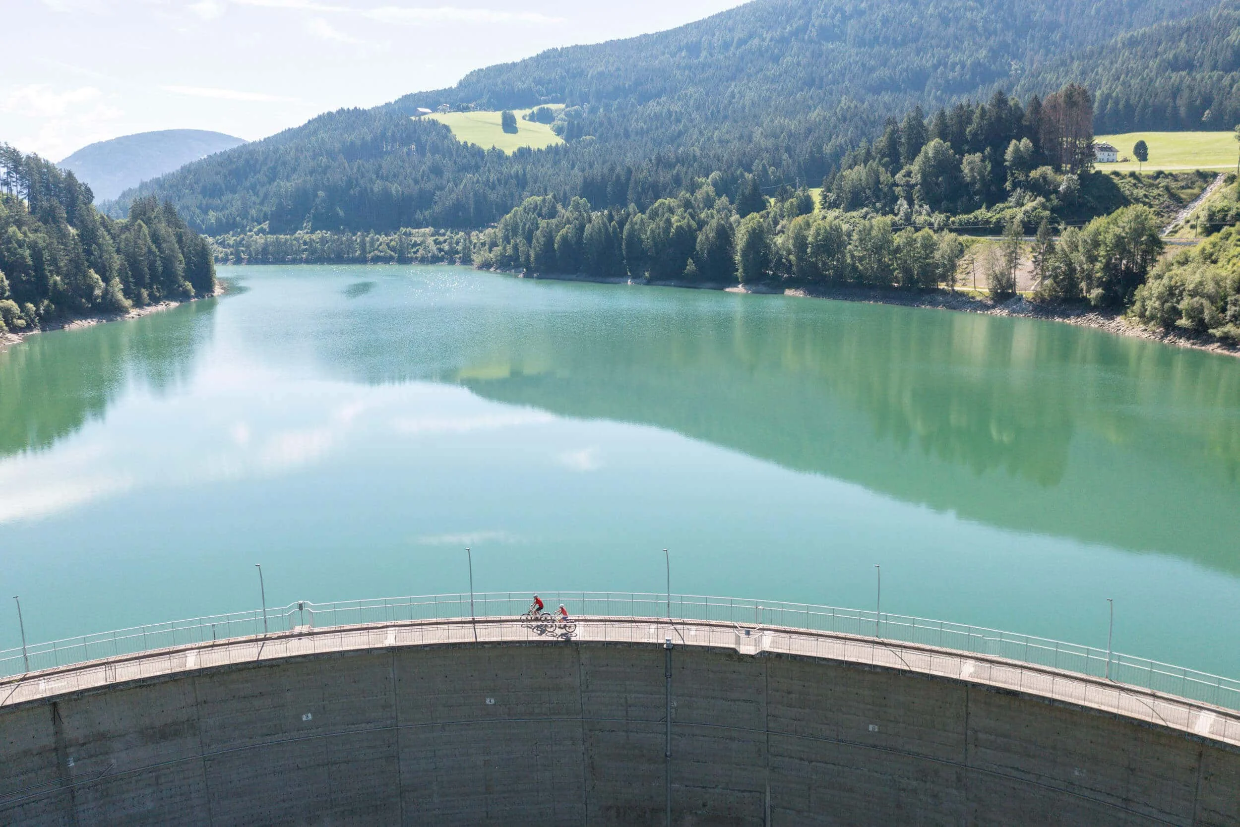 Two cyclists on dam above a reservoir surrounded by forested mountains