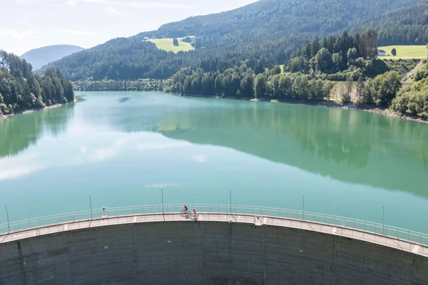 Two cyclists on dam above a reservoir surrounded by forested mountains