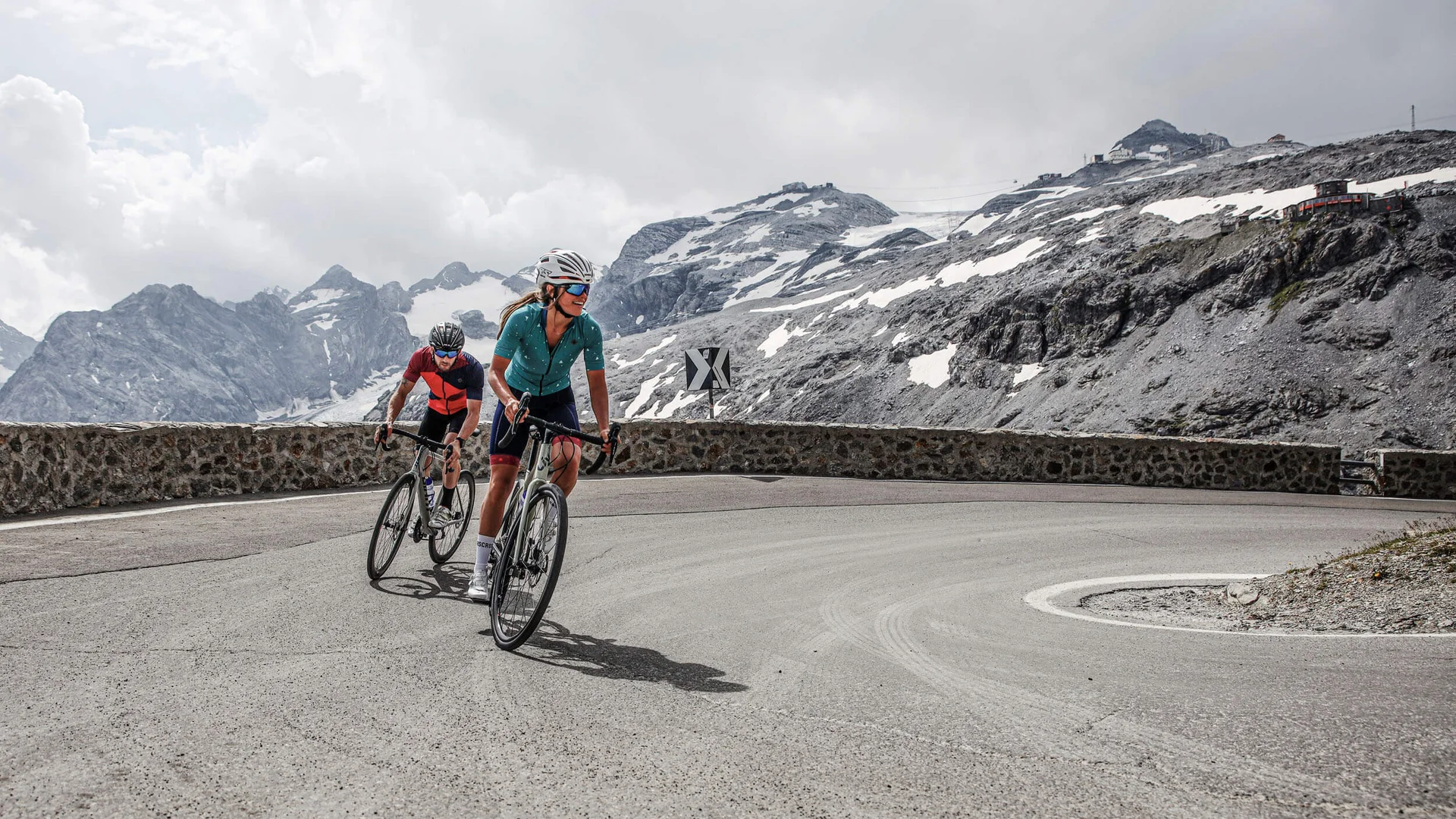 Bike Hotel © Felix Saller Two cyclists riding a mountain road with snow-capped peaks in the background