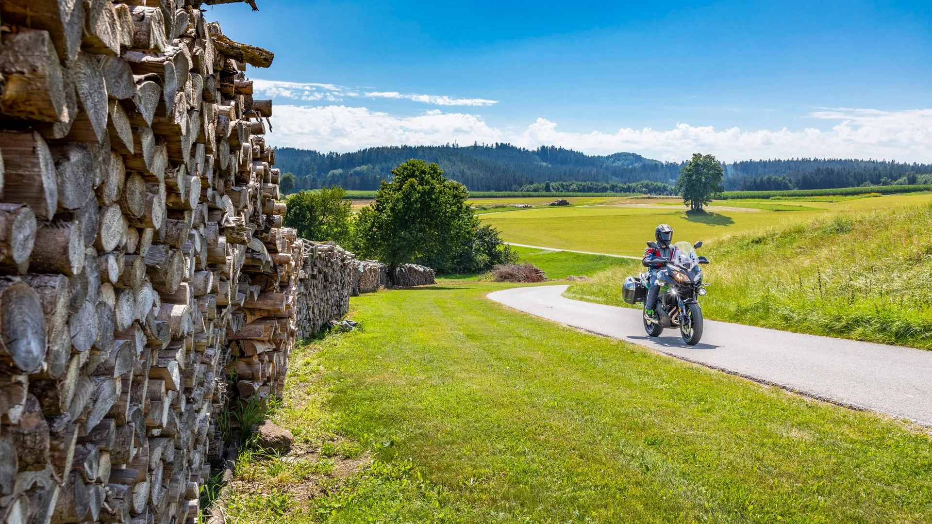 Motorcyclist riding on country road beside large stacked firewood under blue sky