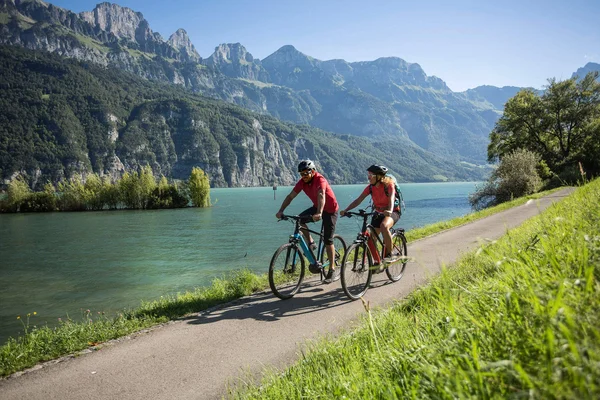 Velontour Two cyclists riding on a path by a lake with mountains in the background
