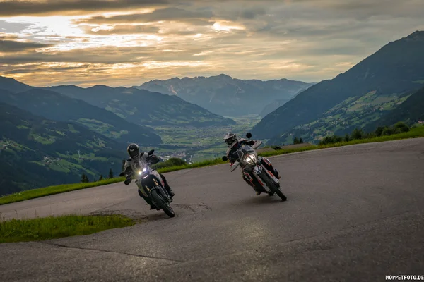Two motorcyclists riding on a winding mountain road with valley and mountains behind