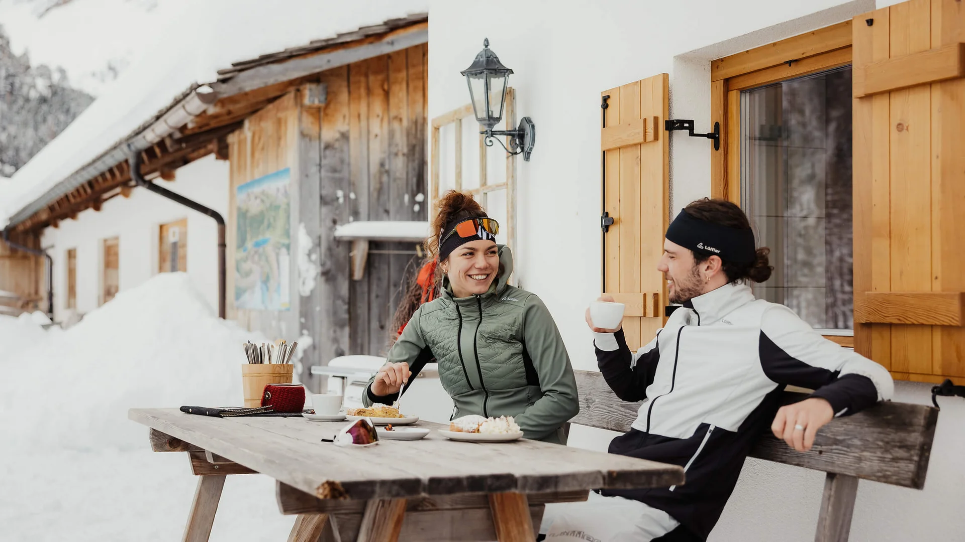 Two people enjoying warm drinks outside a wooden cabin in winter