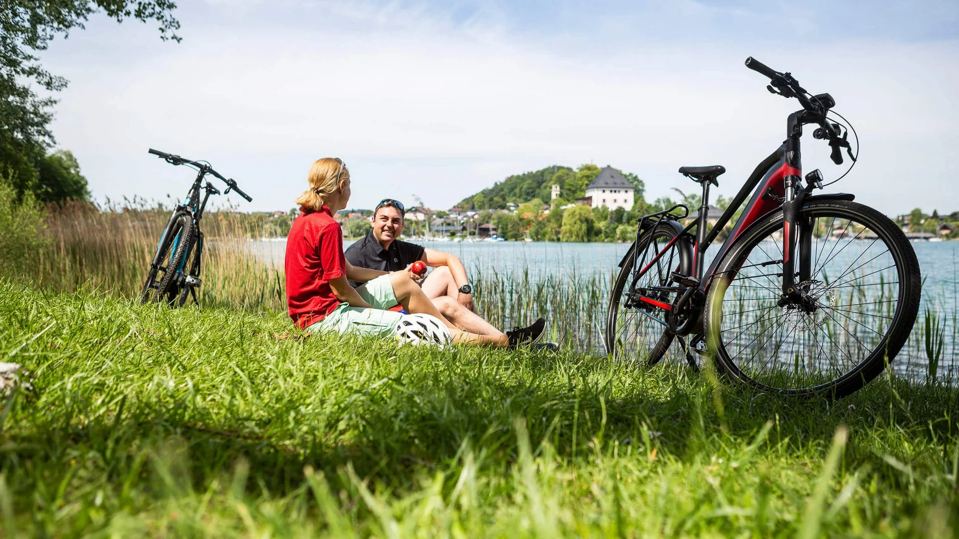 MTS Austria Two cyclists resting on lake shore with their bikes on grass