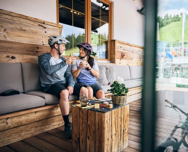 Couple wearing bike helmets drinking coffee in rustic café with mountain bikes outside