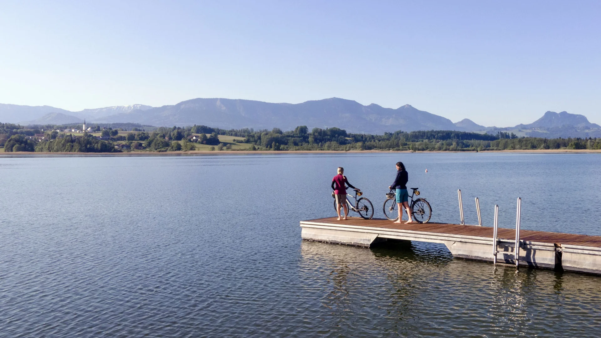 Two people with bicycles on a dock by a lake with mountains in the background