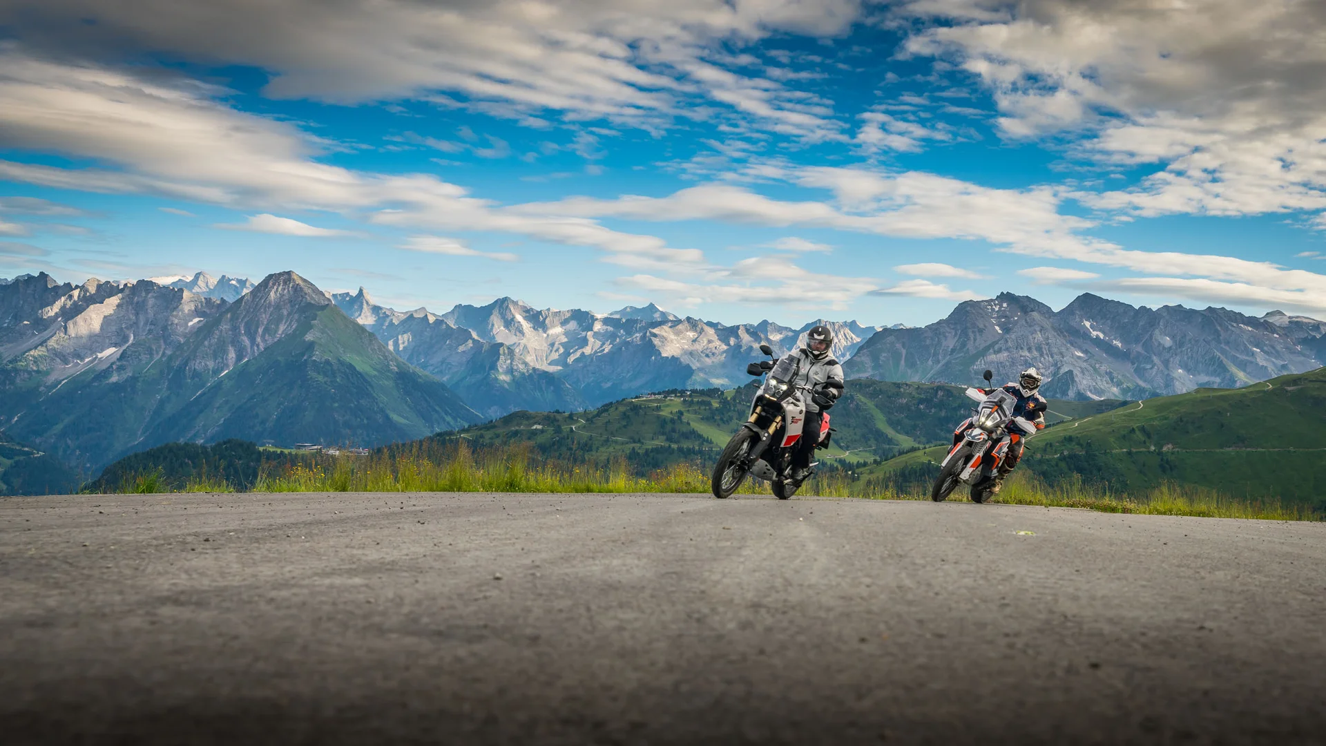 Two motorcyclists riding on mountain road with Alps and cloudy sky