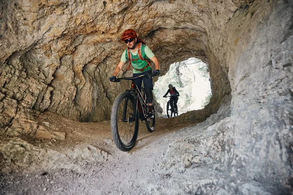 Two mountain bikers riding through a rocky tunnel on a narrow trail