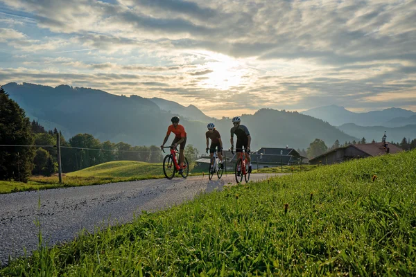Three cyclists riding on rural road with mountains at sunset.