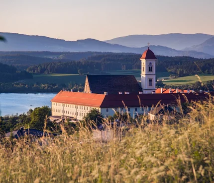 Stift St. Georgen © Elias Jerusalem Klostergebäude mit rotem Dach vor Berg- und Seenlandschaft bei Sonnenuntergang