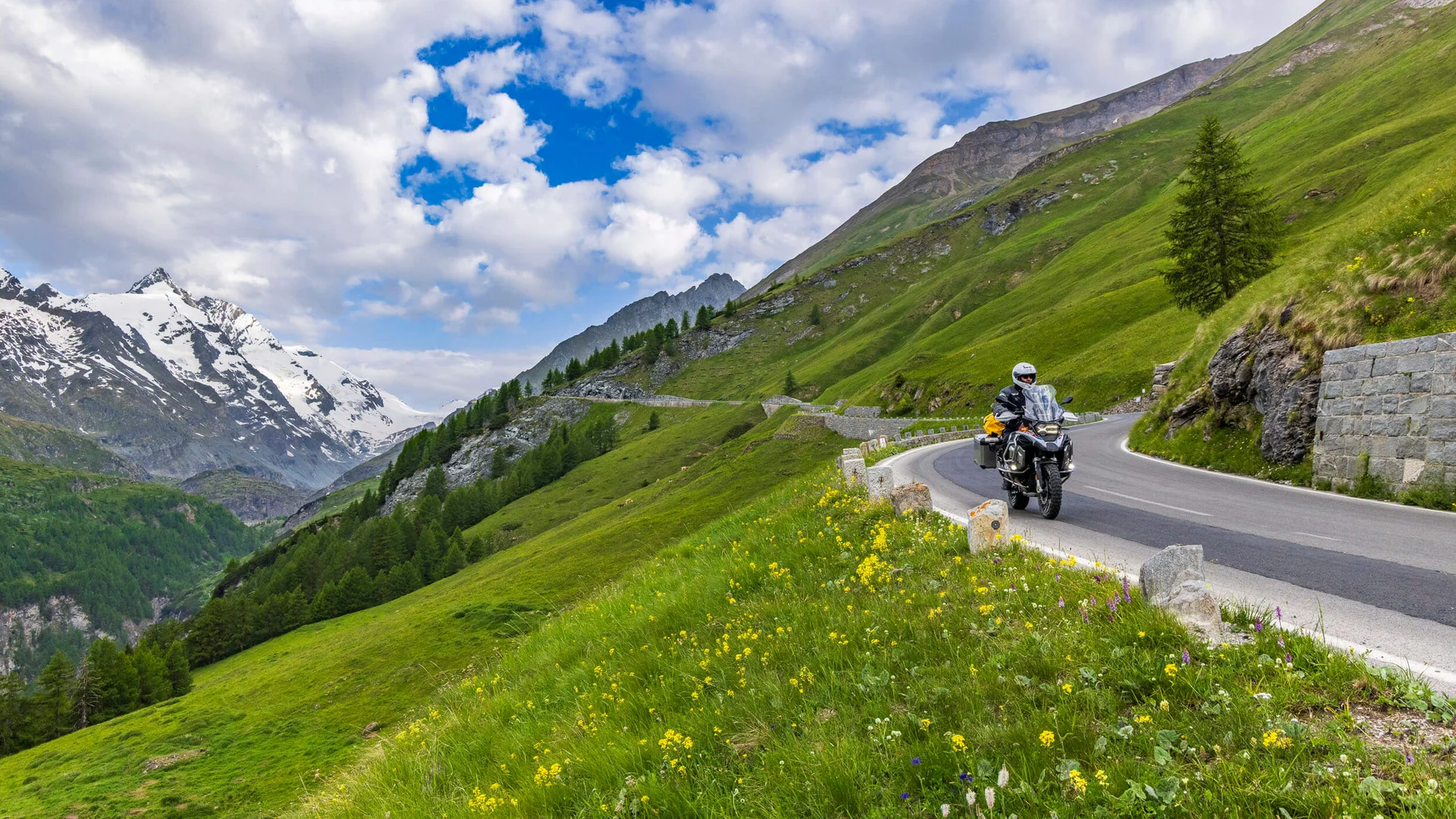 MoHo – Motorbike Hotels © Alexander Seger Motorcyclist riding on winding mountain road with green hills and snowy peaks