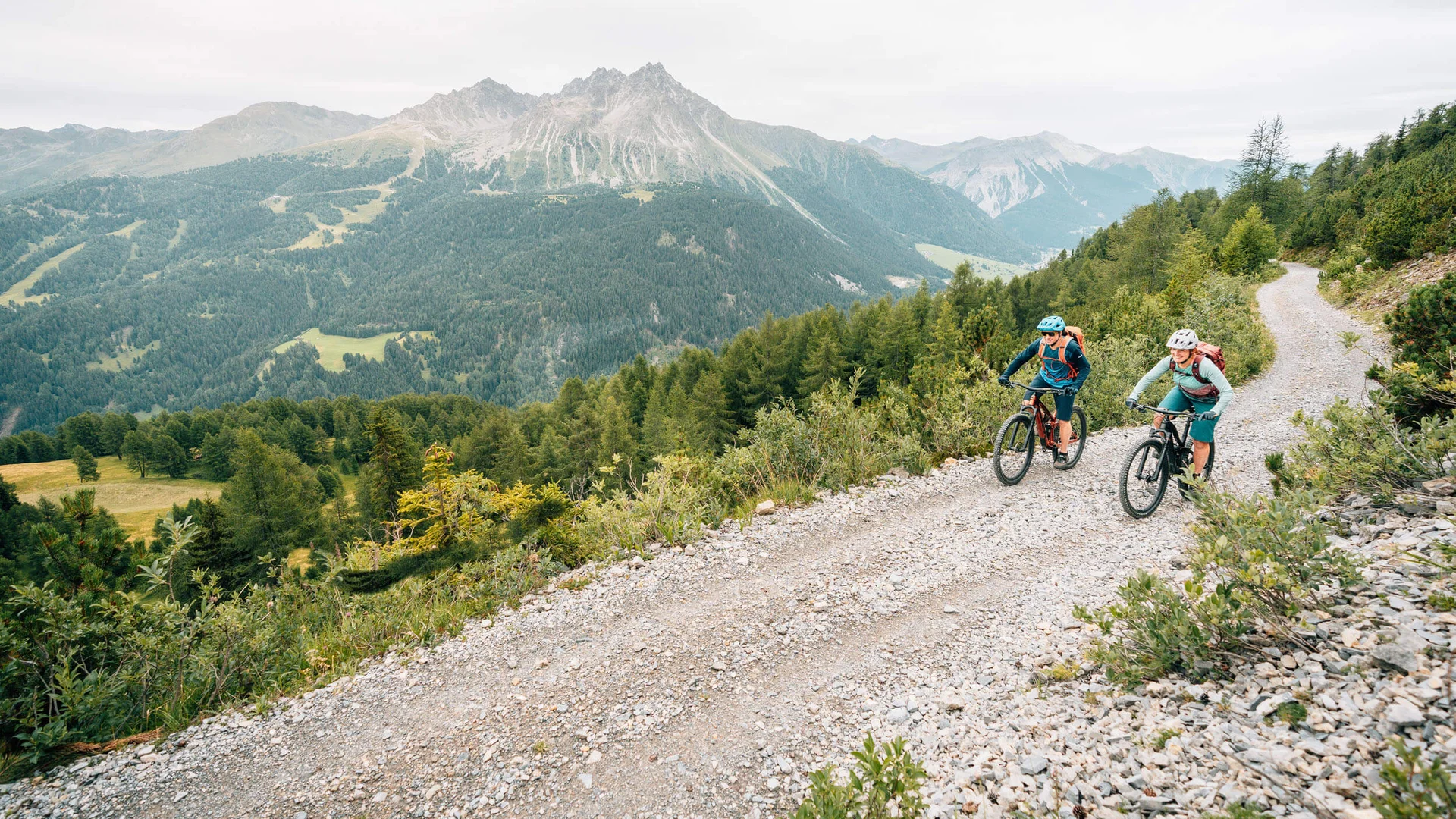 Two cyclists riding on a mountain trail with Alps in the background