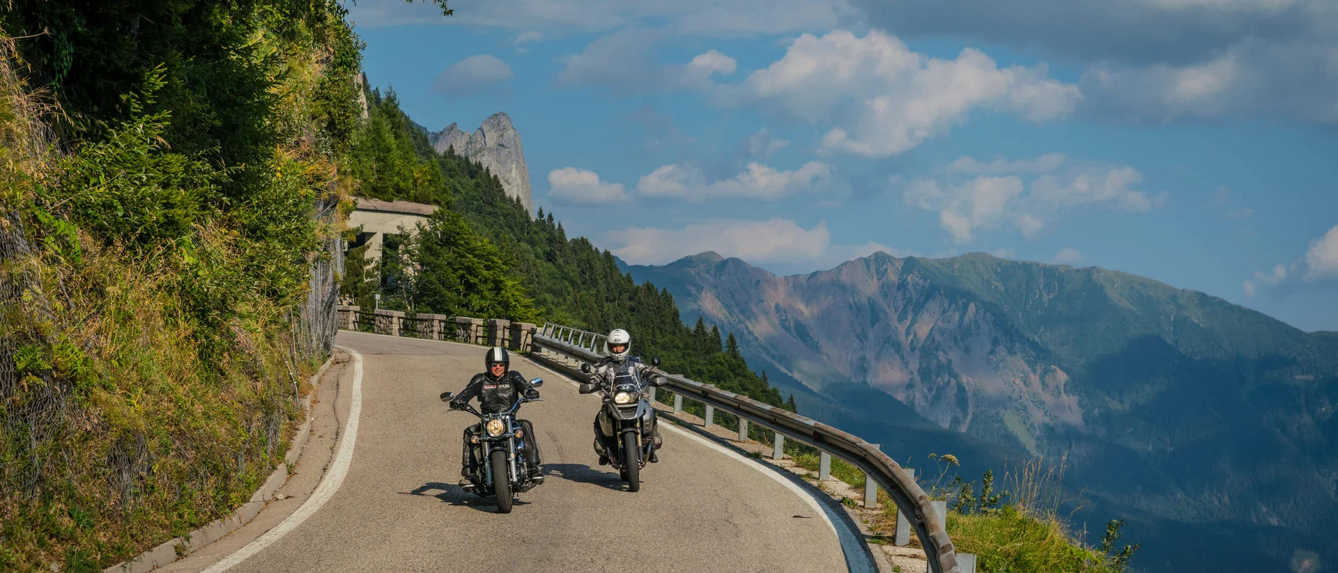 MoHo – Motorbike Hotels © Moppetfoto.de Two motorcyclists riding on a winding mountain road with mountains behind.