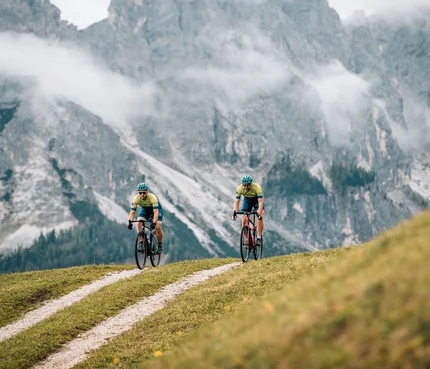 Dolomitenregion Kronplatz © Jens Scheibe Zwei Radfahrer fahren auf einem Bergweg vor nebligen Alpenfelsen
