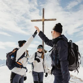 Three hikers giving a high five near a summit cross in snowy mountains